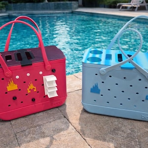 Puede incluir: Dos bolsas de mano coloridas con asas junto a la piscina. Una es rosa vibrante, la otra azul claro. Ambas tienen un gr&aacute;fico de castillo y una forma rectangular con un asa. La bolsa rosa tiene un cierre negro y detalles blancos.