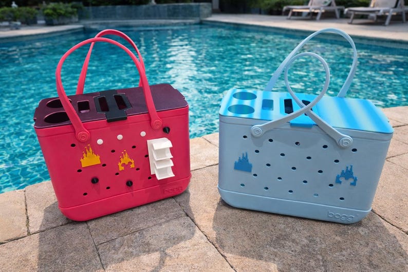 Puede incluir: Dos bolsas de mano coloridas con asas junto a la piscina. Una es rosa vibrante, la otra azul claro. Ambas tienen un gr&aacute;fico de castillo y una forma rectangular con un asa. La bolsa rosa tiene un cierre negro y detalles blancos.