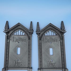 May include: Two gray, gothic-style light switch covers. Each cover has the word "Lumos" above the switch opening and "Nox" below. The covers have an arched top with decorative detailing and are set against a light blue background.