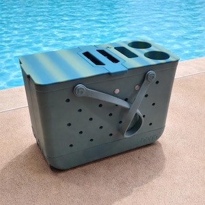 Puede incluir: Una bolsa nevera de color verde azulado y azul con asa y varios portavasos. La nevera tiene forma rectangular con tapa y est&aacute; sobre una superficie de hormig&oacute;n junto a una piscina.