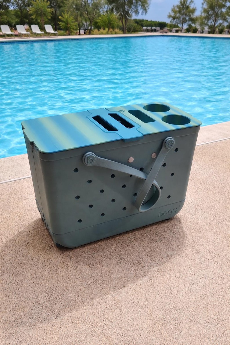 Puede incluir: Una bolsa nevera de color verde azulado y azul con asa y varios portavasos. La nevera tiene forma rectangular con tapa y est&aacute; sobre una superficie de hormig&oacute;n junto a una piscina.