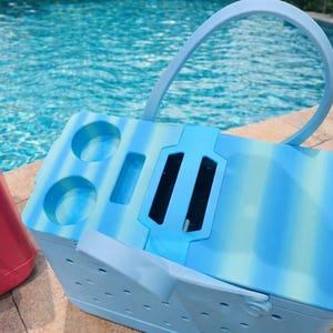 Puede incluir: Una nevera a rayas azules y blancas con asa y portavasos. La nevera est&aacute; al lado de una nevera roja. El fondo es una piscina.