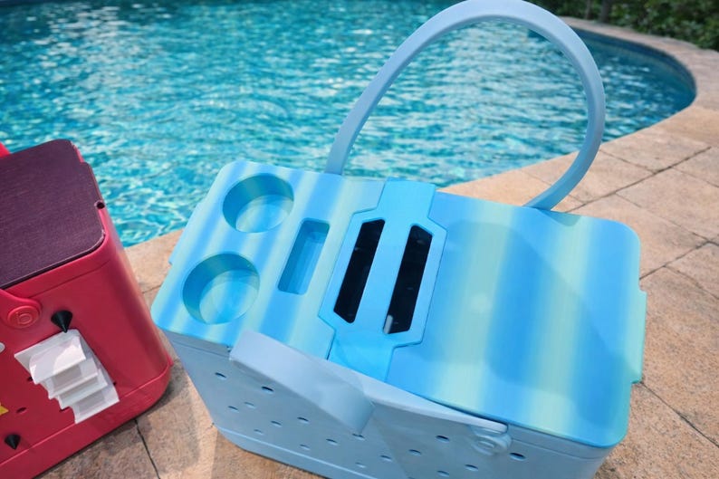 Puede incluir: Una nevera a rayas azules y blancas con asa y portavasos. La nevera est&aacute; al lado de una nevera roja. El fondo es una piscina.