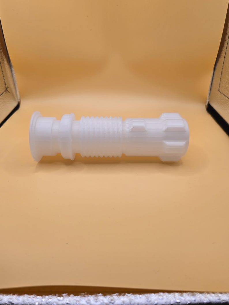 Pu&ograve; includere: Un'elsa di spada laser bianca stampata in 3D. L'elsa presenta un design cilindrico con sezioni a coste e strutturate e un pomello dettagliato. Il design generale ricorda un'arma fantascientifica.