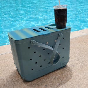 Puede incluir: Una bolsa nevera de color verde azulado y azul con una bebida en un vaso transparente con pajita. La nevera tiene un asa y varios agujeros. Est&aacute; sobre una superficie de hormig&oacute;n junto a una piscina.