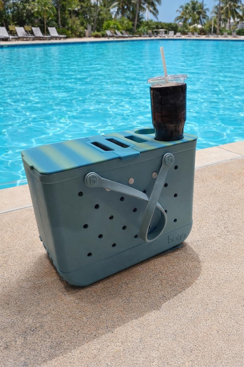 Puede incluir: Una bolsa nevera de color verde azulado y azul con una bebida en un vaso transparente con pajita. La nevera tiene un asa y varios agujeros. Est&aacute; sobre una superficie de hormig&oacute;n junto a una piscina.