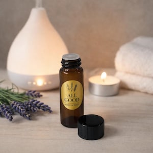 May include: A brown glass bottle with a black cap and a gold label that reads "ALL GOOD HOME & BODY" sits next to a white essential oil diffuser. A lit candle and lavender sprigs are also in the image.