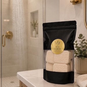 May include: A black stand-up pouch with a clear window containing several square white soap bars embossed with a floral design. A circular gold label on the front features the text ALL GOOD HOME & BODY with a small botanical illustration above it.