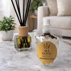 May include: A clear glass reed diffuser with black reeds and a wooden collar, next to a refill pouch. The pouch has a gold label with the text "ALL GOOD HOME & BODY Reed Diffuser Refill". The items are on a marble surface.