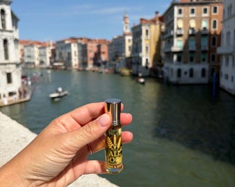 May include: A small clear glass roller bottle with a silver cap containing a golden liquid and botanical elements. The label features the text ALL GOOD HOME & BODY with a wheat stalk graphic. The bottle is held against a canal and city backdrop.