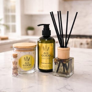 May include: A collection of home and body products, including a candle in a glass jar with a wooden lid, a small bottle, a hand soap bottle, and a reed diffuser. The products have gold labels with lavender sprigs and the text "ALL GOOD HOME & BODY".