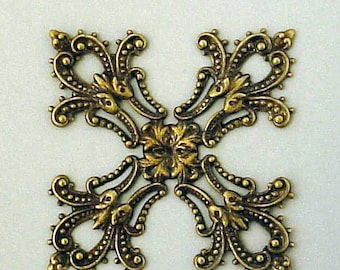May include: A decorative brass filigree cross-shaped embellishment. The ornate design features floral and scrollwork details, with a central flower and beaded accents. The metal has a warm, antique gold finish.
