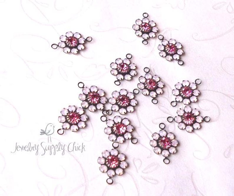 May include: A collection of floral-shaped jewelry connectors. Each connector features a central pink gemstone surrounded by white opalescent stones. The connectors are connected by small loops and are set against a white background.