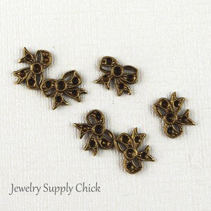May include: Five antique gold-toned metal bow embellishments with a decorative design. The bows are scattered on a white surface.
