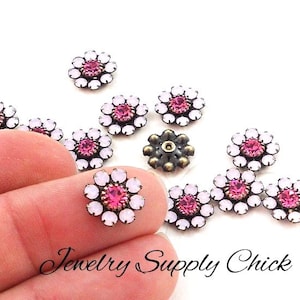 May include: A collection of flower-shaped embellishments with pink and white crystals. One is held between fingers. The embellishments have a dark metal base. The text "Jewelry Supply Chick" is at the bottom.
