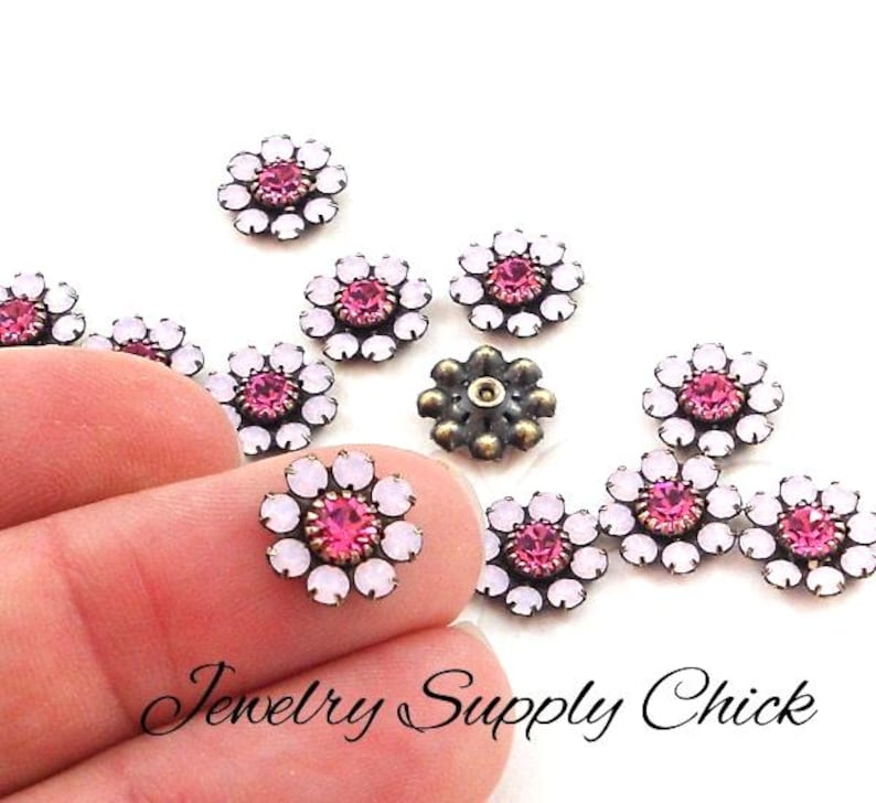 May include: A collection of flower-shaped embellishments with pink and white crystals. One is held between fingers. The embellishments have a dark metal base. The text "Jewelry Supply Chick" is at the bottom.