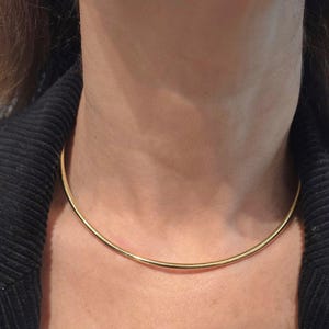 Peut inclure: Un collier ras du cou minimaliste de couleur or. Le collier est une fine bande incurvée qui se porte près du cou. Le bijou est présenté sur un fond neutre.