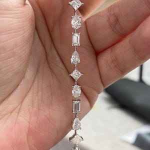 May include: A silver-toned tennis bracelet featuring a repeating pattern of clear, faceted stones in various shapes, including pear, princess, oval, and emerald cuts, linked together in a vertical line with a polished metallic setting.