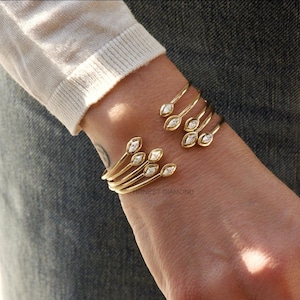 Könnte beinhalten: Goldfarbenes Armreif-Armband mit mehreren gebogenen Bändern, die jeweils mit marquiseförmigen Edelsteinen besetzt sind. Das Armband umschließt das Handgelenk und zeigt ein zartes und elegantes Design. Der Text "HONEST DIAMOND" ist sichtbar.