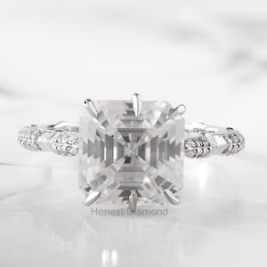 Peut inclure: Gros plan sur une bague de fiançailles en diamant. La bague présente un grand diamant de taille carrée serti dans un sertissage à griffes. Le groupe est orné de petites pierres d'accent et de motifs complexes. Le texte "Honest Diamond" est visible.