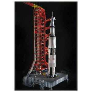 May include: A detailed model of a Saturn V rocket on a launch pad, with a red and yellow gantry. The rocket is white with black accents and the letters "USA" on the side. The model is set against a black background.