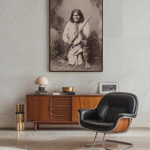 Op de afbeelding: Een ingelijste sepia-foto van een indiaanse man met een geweer. De foto hangt boven een houten kast in mid-century modern stijl met lades en een zwarte leren fauteuil.