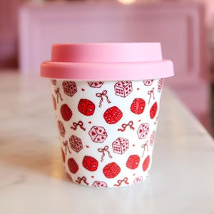 May include: A white ceramic travel mug with a pink silicone lid, featuring a repeating pattern of red and pink dice and small bows. The dice are decorated with tiny heart symbols on each face, creating a consistent graphic design across the cup.