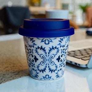 May include: A reusable travel cup featuring a white ceramic body decorated with a repeating blue floral pattern and a solid dark blue silicone lid. The cup has a tapered cylindrical shape and is designed for holding hot or cold beverages.