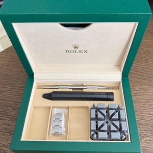 Peut inclure: Une boîte verte avec le logo Rolex sur le couvercle intérieur, contenant des outils de réparation de montres. Les outils comprennent un outil argenté, un outil noir, un bracelet de montre et un support de montre gris.