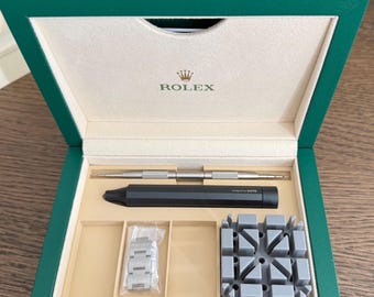 May include: A green box with the Rolex logo on the inside lid, containing watch repair tools. The tools include a silver tool, a black tool, a watch band, and a gray watch holder.