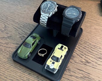 May include: A black watch display stand holding two watches with silver and black bands, a gold ring, and two toy cars, one green and one yellow. The stand has a black velvet surface for the watches.
