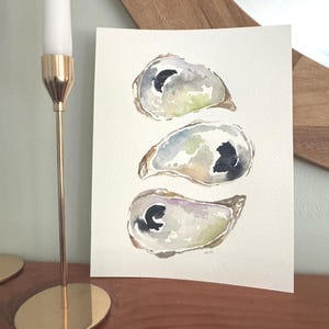 May include: A watercolor painting on textured paper featuring three oyster shells arranged vertically. The shells are rendered in soft washes of gray, blue, green, and brown, with dark, concentrated ink spots representing the oyster meat.
