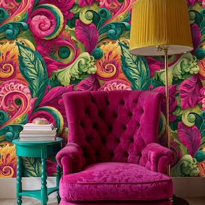 May include: A tufted magenta armchair sits before a wall covered in vibrant wallpaper featuring swirling pink, green and yellow floral patterns. A teal side table holds a stack of books, and a floor lamp with a mustard yellow shade stands nearby.