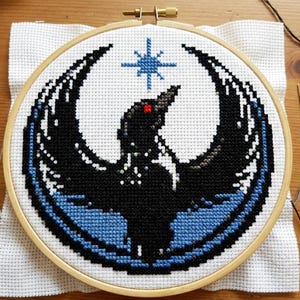 Puede incluir: Bordado de punto de cruz de un pájaro negro con las alas extendidas, un ojo rojo y una estrella azul encima. El diseño está sobre un fondo blanco dentro de un bastidor de bordado de madera. El pájaro está rodeado por un borde azul.