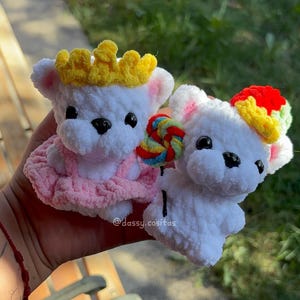 May include: Two white crochet teddy bears with black eyes and noses. One wears a pink dress and a yellow crown, holding a lollipop. The other has a colourful hat and a lollipop. The bears are handmade.