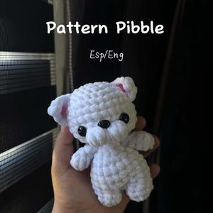 Puede incluir: Un oso de ganchillo blanco hecho a mano con ojos de botón negros y orejas internas rosas. El oso es pequeño y suave, con costuras visibles. La imagen incluye el texto "Pattern Pibble Esp/Eng".