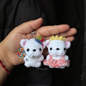 Peut inclure: Deux porte-clés ours blancs au crochet. L'un tient une sucette et porte un chapeau, l'autre porte une robe rose et une couronne. Les porte-clés sont faits à la main et présentent de petits détails.
