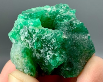 May include: A raw, unpolished emerald crystal specimen featuring a vibrant, deep green color with a translucent, crystalline texture and jagged, uneven edges. The mineral displays natural facets and internal inclusions throughout its structure.