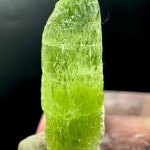 May include: A vibrant, translucent green peridot crystal, standing upright on a small, light-colored rock base. The crystal's surface is textured with visible striations and a natural, irregular shape. The background is dark, emphasizing the crystal's color and form.
