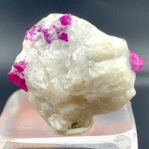 May include: A raw, uncut mineral specimen featuring a matrix of creamy white stone with vibrant, hot pink ruby crystals embedded throughout. The rough texture and color contrast create a striking display.
