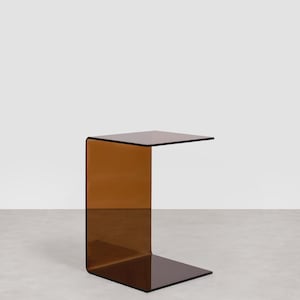 May include: A modern, C-shaped side table in a warm amber hue. The table is constructed from a single sheet of transparent acrylic, featuring a square top and base connected by a curved side. The design is minimalist and contemporary.