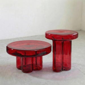 May include: Two red glass side tables. One is a low, round table with a three-legged base. The other is a taller, cylindrical table. Both tables have a glossy finish and are made of transparent red glass. The tables are suitable for a living room or bedroom.