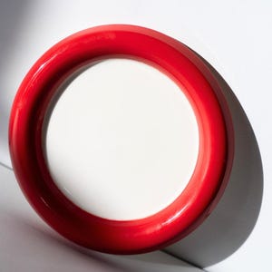 May include: A circular plate with a red rim and a white interior. The plate is set against a white backdrop, with a shadow to the right. The plate's glossy surface reflects the light.