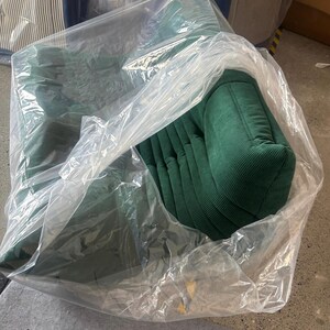 May include: A green corduroy armchair, partially visible through a clear plastic wrapping. The chair has a rounded back and armrests, with vertical ribbed detailing. Other chairs are visible in the background.