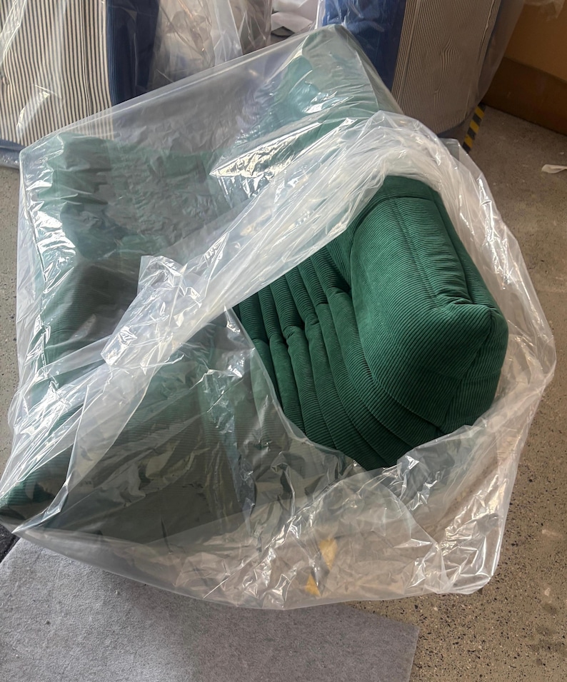 May include: A green corduroy armchair, partially visible through a clear plastic wrapping. The chair has a rounded back and armrests, with vertical ribbed detailing. Other chairs are visible in the background.