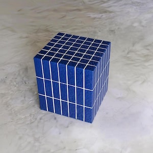 May include: A blue cube with a grid pattern of white lines. The cube is made of rectangular blocks, each outlined with white lines. The cube is sitting on a gray surface.