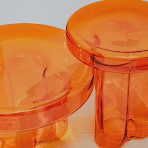 Puede incluir: Dos mesas auxiliares naranjas translúcidas. Cada mesa tiene una parte superior redonda y una base con un diseño curvo único. Las mesas están hechas de un material brillante y proyectan sombras sobre la superficie inferior.