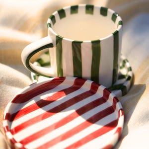 Puede incluir: Un juego de taza y platillo de cerámica blanca con rayas verticales verdes y un plato a juego a rayas rojas y blancas. La taza tiene un asa y está llena de un líquido claro. El juego está sobre una tela clara.