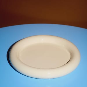 May include: A cream-coloured, round ceramic dish with a raised, rounded rim. The dish sits on a bright blue surface, with a brown background. The dish is a simple, minimalist design.