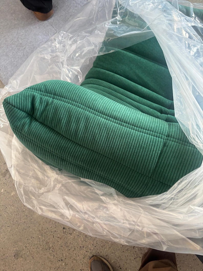 May include: A green corduroy chair cushion, partially encased in clear plastic. The cushion has a ribbed texture and a rounded shape. The color is a deep, forest green. The chair appears to be new and unused.